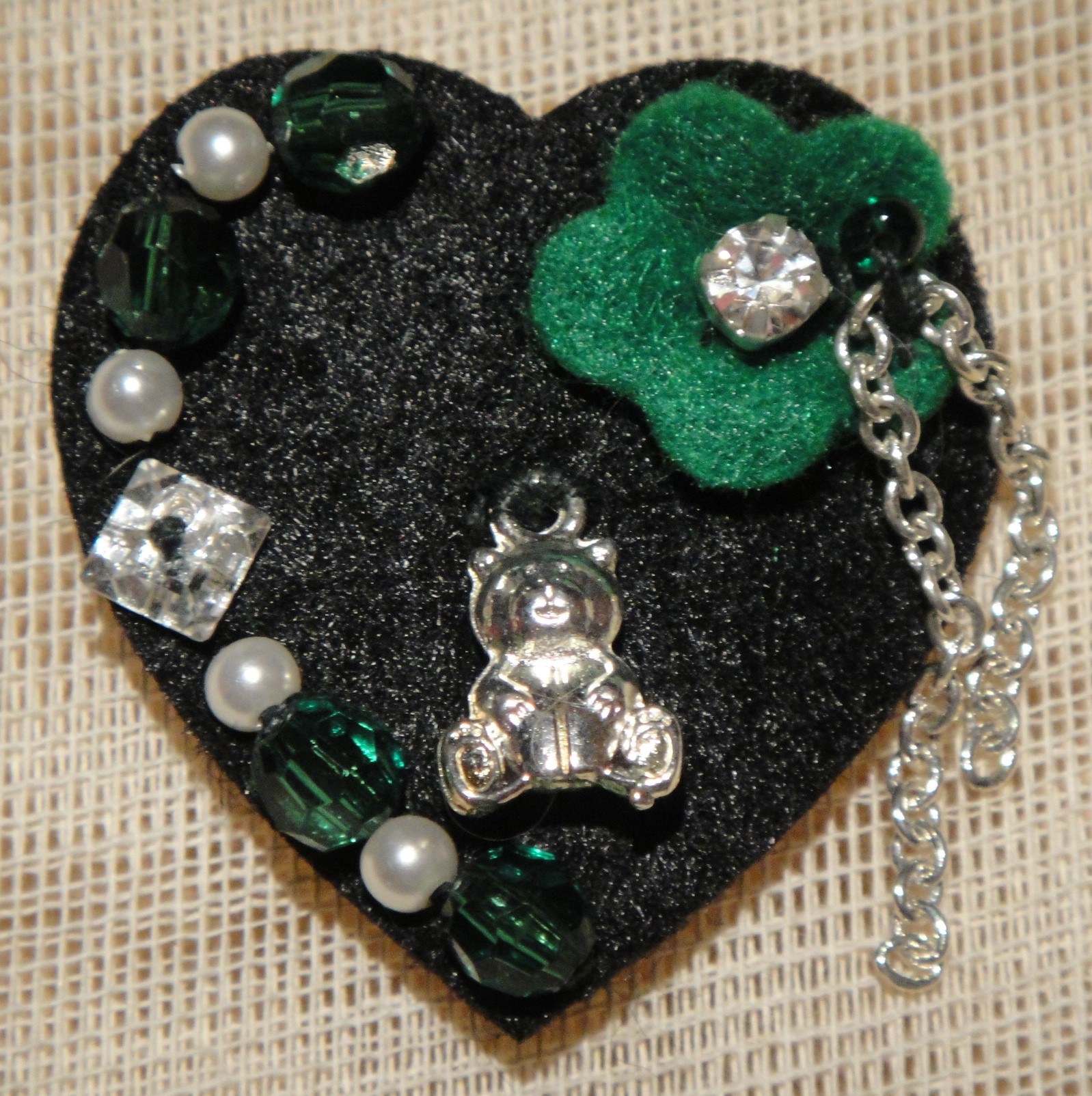 BlackFabricHeart-Green-Flower. Χειροποίητη καρφίτσα Cosa Nostra από τσόχινη βάση-καρδιά κεντημένη στο χέρι με τσόχινο πράσινο λουλούδι, αλυσίδα νίκελ, χάντρες, μεταλλικό αρκουδάκι και Swarovski. Διαστάσεις: 4cm x 3cm.